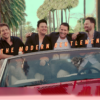 Four men sit atop the headrest of a bench seat in a red convertible. Tall palm trees line the street behind them.