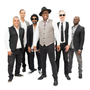 A band of six stand together, each wearing a white button up shirt with a vest or jacket and black pants. Billy stands at the center wearing a hat and floral tie.