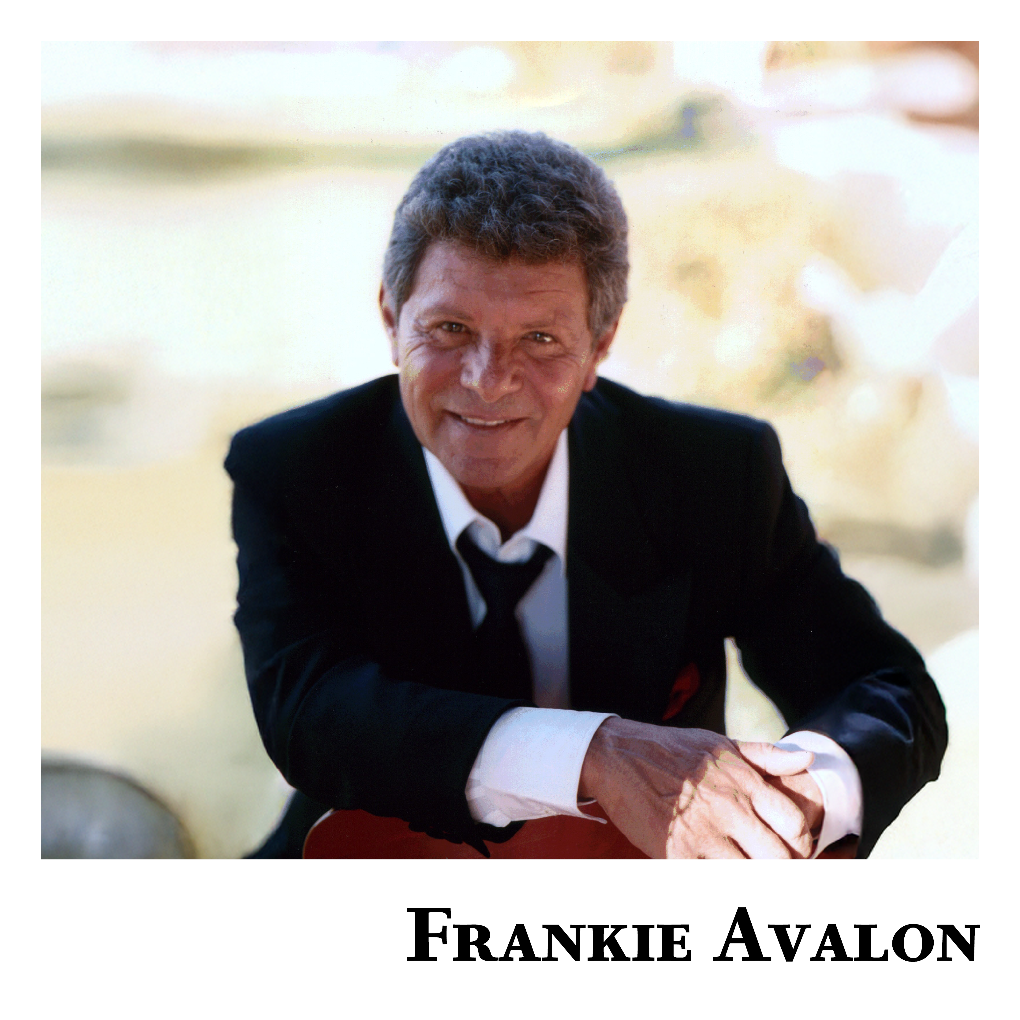 “Singer Frankie Avalon, dressed in a black suit, white shirt, and loosened black tie, smiles warmly while leaning forward with one arm resting on a chair. The soft, blurred background gives a sunlit, golden glow. His name ‘Frankie Avalon’ is written in bold black letters at the bottom of the image.”