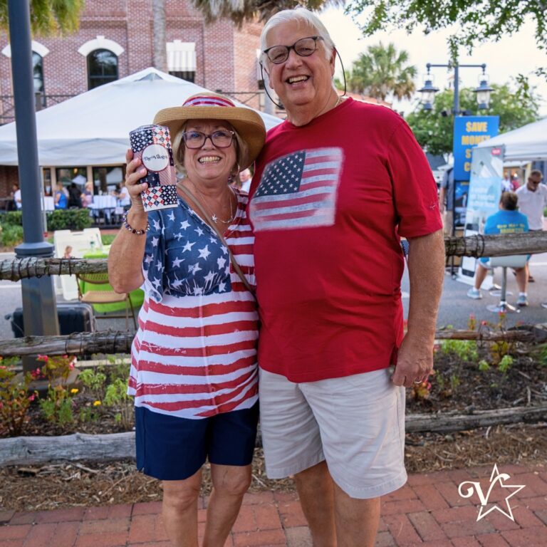 Smiling couple dressed in American flag-themed outfits, holding a drink and posing together at an outdoor patriotic event with vendor tents in the background.