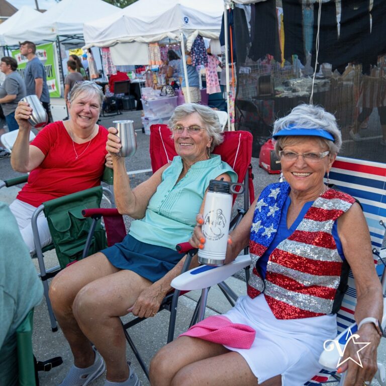 Three women sitting in folding chairs at an outdoor market, smiling and holding drinks, with vendor tents and booths in the background.