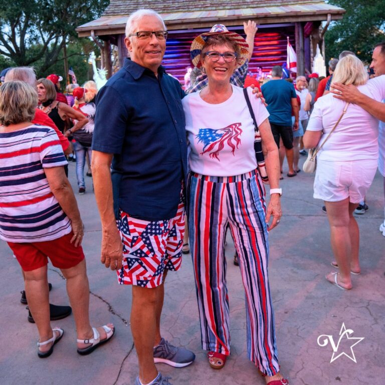 Smiling couple wearing patriotic red, white, and blue outfits standing in front of a lit stage, surrounded by a crowd at an outdoor Independence Day celebration.