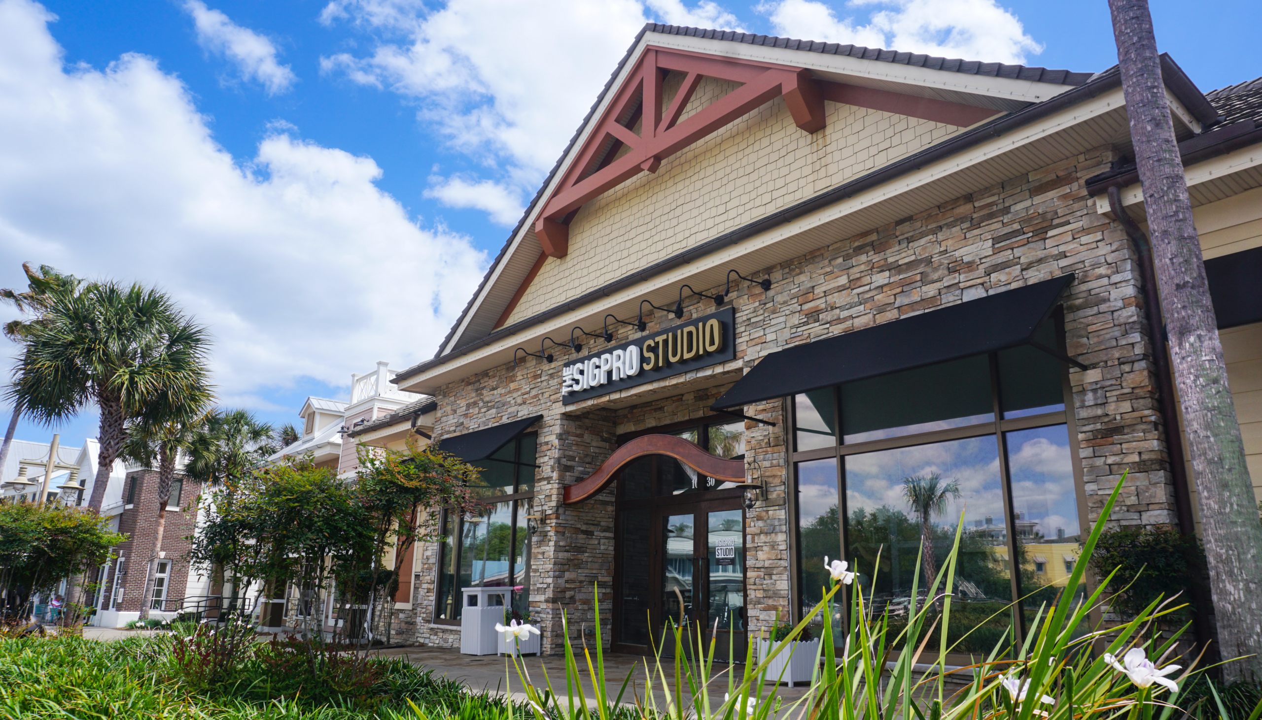 Exterior view of a stone-and-stucco building with a peaked roof and black awnings, displaying a sign reading “The SIGPRO Studio.” Palm trees, landscaped greenery, and a sidewalk are in the foreground under a bright blue sky with scattered clouds.