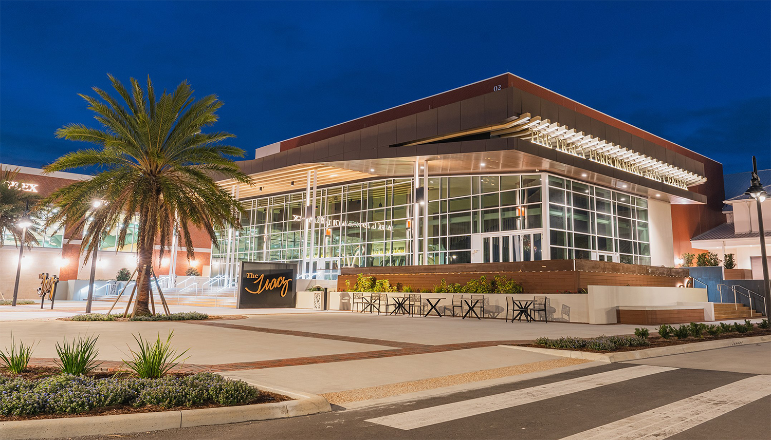 An exterior photo of The Tracy Performing Arts Center. Palm trees and landscaping are in the foreground with the building well lit under a night sky night.
