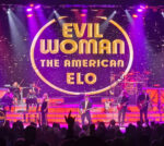 A live concert scene featuring a band performing onstage in front of a large illuminated screen displaying the a gold circle with text “Evil Woman: The American ELO” and stars filling the screen. Multiple performers are visible across the stage playing instruments including guitars, keyboard, and string instruments. The stage is lit with purple, pink, and orange lights. Audience silhouettes appear in the foreground with raised hands.