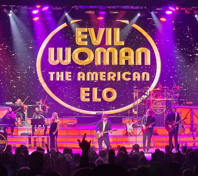 A live concert scene featuring a band performing onstage in front of a large illuminated screen displaying the a gold circle with text “Evil Woman: The American ELO” and stars filling the screen. Multiple performers are visible across the stage playing instruments including guitars, keyboard, and string instruments. The stage is lit with purple, pink, and orange lights. Audience silhouettes appear in the foreground with raised hands.
