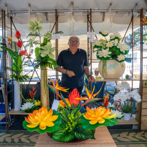 A man poses in his vendor booth, surrounded by his realistic floral sculptures.
