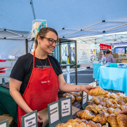 A baker wearing a red apron is standing behind a table filled with pastries. She is seen passing her customer a croissant.
