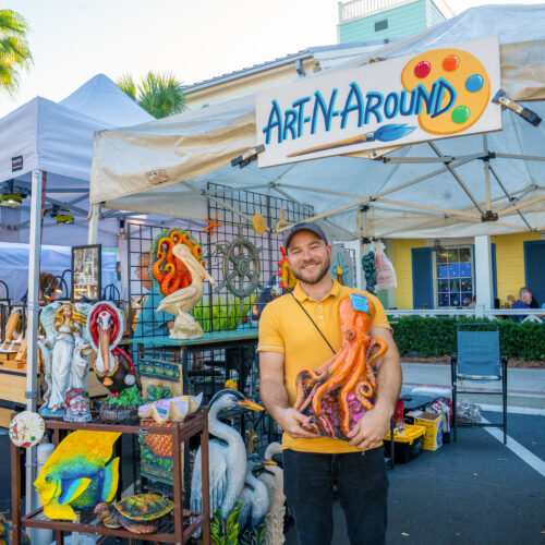 A man holding a bright orange octopus sculpture in front of a tent with a sign that reads "Art N' Around"