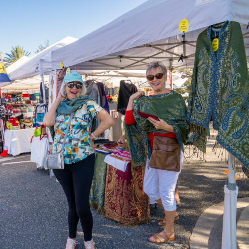 Two women are trying on multipurpose scarves at an artisan's booth. The woman on the left is posing for the camera with her hand on her hip and smiling.