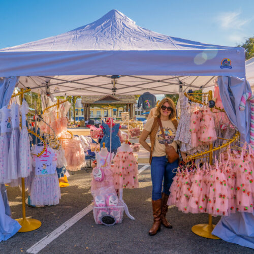 A woman is standing at the front of her tent, filled with little girls' clothing and accessories for sale.