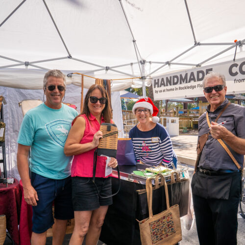Happy customers pose with their purchase under a handmade bag vendor's tent. The vendor is wearing a Santa hat.