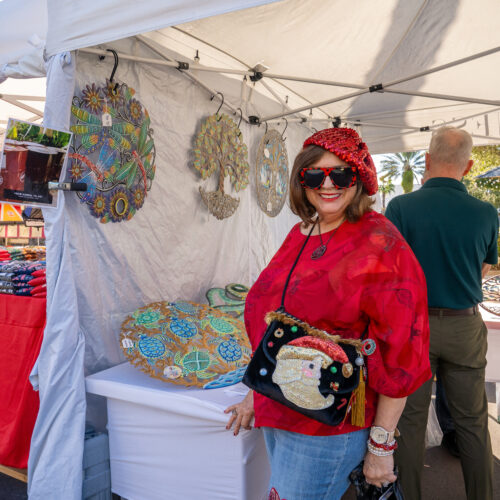 A woman dressed in a red sweater, matching beret, and a Santa purse, poses next to a vendor's booth.