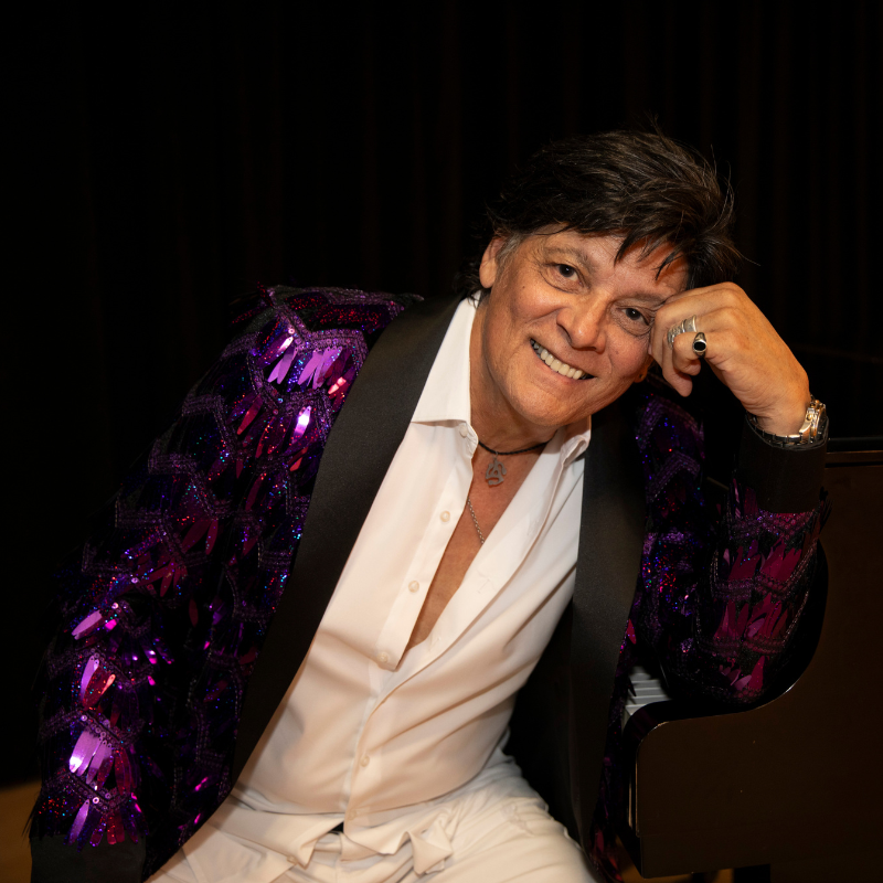 Promotional poster for Mannequin, The Roy Michaels Experience at Playhouse Performances. Pictured is a man sitting on a chair and resting his head on his fist. He is wearing white with a purple sequin jacket.