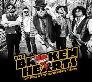 Promotional poster for The Broken Hearts – Tom Petty & The Heartbreakers Tribute. 5 band members are pictured in black and white standing on a bridge surrounded by trees. They are wearing sunglasses and 2 of the band members are carrying guitars. At the bottom of the image is the title written in black and lined with yellow. The O in broken is the shape of a heart with a guitar arrow through it that reads "In Petty We Trust."