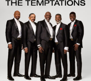 The five current members of The Temptations are wearing black tuxedos, with red flowers on their lapels, and pointing at the viewer. They are standing in front of a light gray background. The text at the top of the image in black, all capital lettering, says "THE TEMPTATIONS."