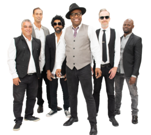 A band of six stand together, each wearing a white button up shirt with a vest or jacket and black pants. Billy stands at the center wearing a hat and floral tie.