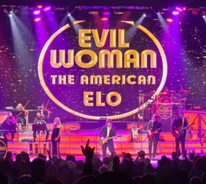A live concert scene featuring a band performing onstage in front of a large illuminated screen displaying the a gold circle with text “Evil Woman: The American ELO” and stars filling the screen. Multiple performers are visible across the stage playing instruments including guitars, keyboard, and string instruments. The stage is lit with purple, pink, and orange lights. Audience silhouettes appear in the foreground with raised hands.