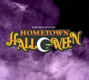"Brownwood Hometown Halloween" Orange letters with the O in Halloween white and in the shape of a crescent moon with green bats flying through. The background contains purple smoke/fog.