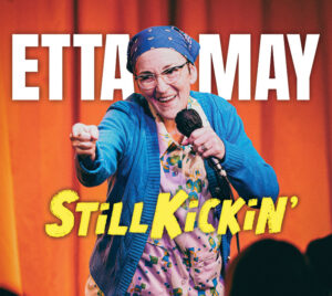 Promotional image reading “Etta May – Still Kickin’,” showing a performer on a warmly lit stage, holding a microphone and gesturing toward the audience.