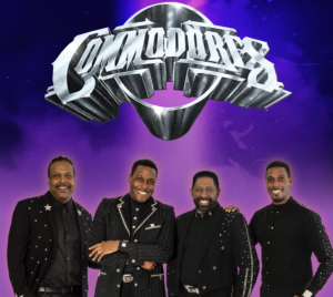 Four members of The Commodores stand smiling in front of a purple background with the band’s large metallic logo above them. They are dressed in coordinated black outfits with sparkling accents.
