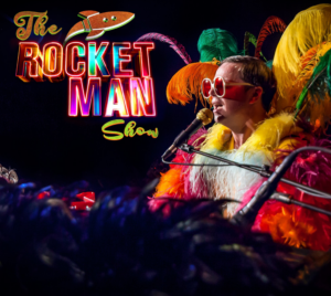 Promotional poster for The Rocket Man Show. A performer wearing many bright feathers is pictured playing a piano while singing. In the top left corner is a colorful logo including a rocket and text that reads: "The Rocket Man Show."