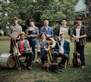 Jazz band Tuba Skinny posing outdoors with their instruments, including banjo, trumpet, clarinet, trombone, guitar, tuba, washboard, and drums. Two members are seated with a dog resting at their feet while the rest stand behind them in a casual group photo.