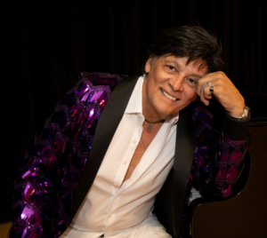 Promotional poster for Mannequin, The Roy Michaels Experience at Playhouse Performances. Pictured is a man sitting on a chair and resting his head on his fist. He is wearing white with a purple sequin jacket.