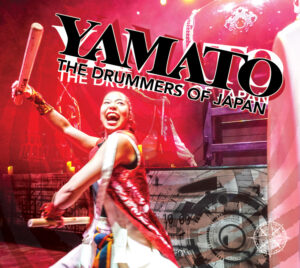 A promotional graphic shows a stage performer holding drumsticks during a live percussion performance, set against dramatic red lighting and layered drum imagery. Large stylized text reads “YAMATO: The Drummers of Japan.” A white compass is displayed in the bottom right corner.