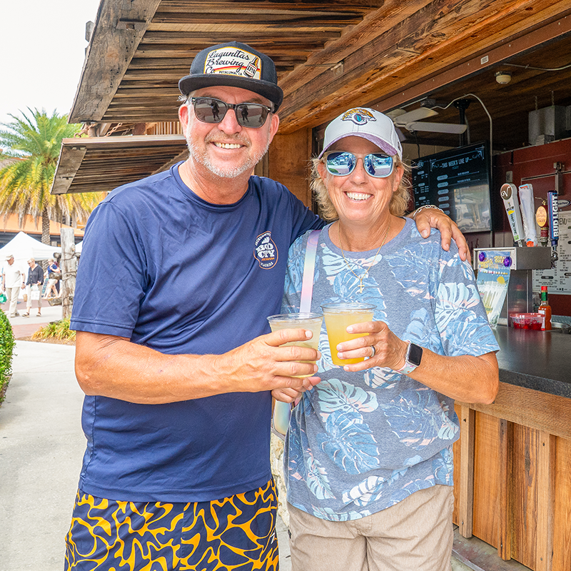 A couple clinks glasses in front of a bar hut.