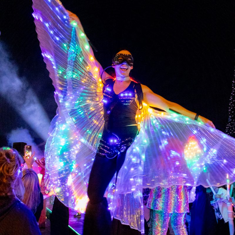 Brightly Light up women stilt walker showing off her light up wings as she walks through the crowd at electric light party