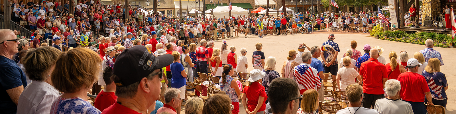 Large crowd gathered in a town square wearing red, white, and blue, facing a stage during a patriotic community celebration with American flags and live entertainment.