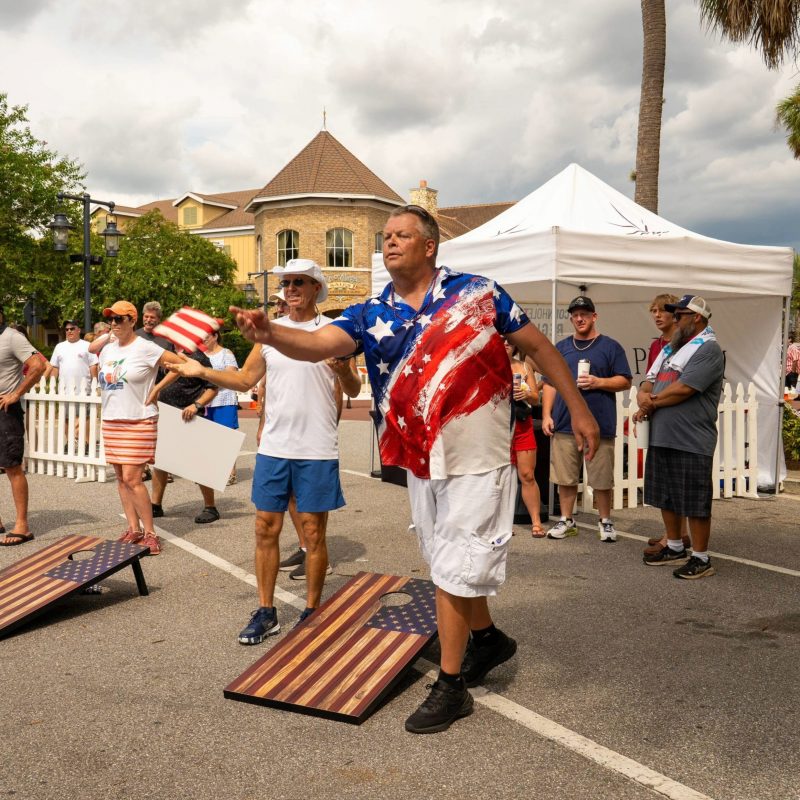 Man wearing a red, white, and blue shirt tossing a bean bag during a cornhole game, with spectators and vendor tents in the background at an outdoor patriotic event.